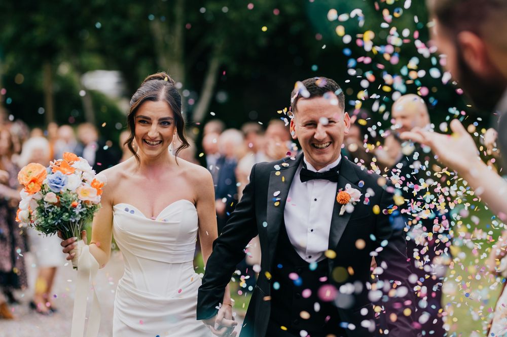 Wedding guests celebrate by throwing white confetti during an outdoor celebration in a garden setting.