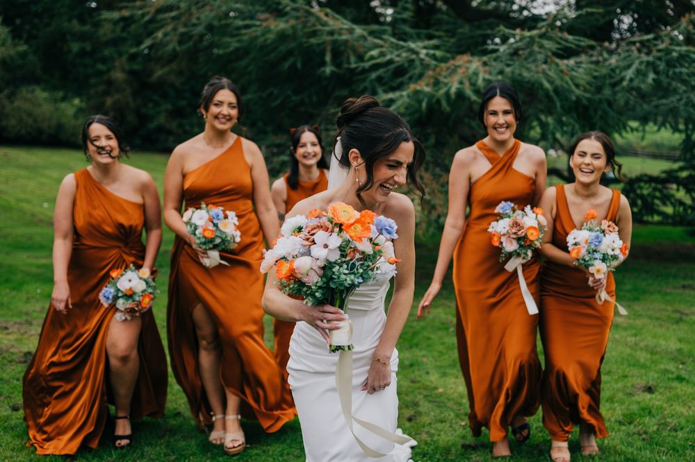 Wedding party in burnt orange bridesmaid dresses and white bridal gown pose with bouquets on green lawn.