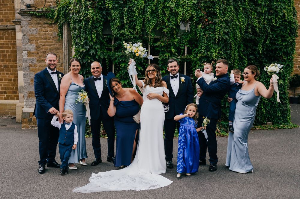 Wedding party spontaneously reacts with laughter together while lined up against brick wall with greenery.