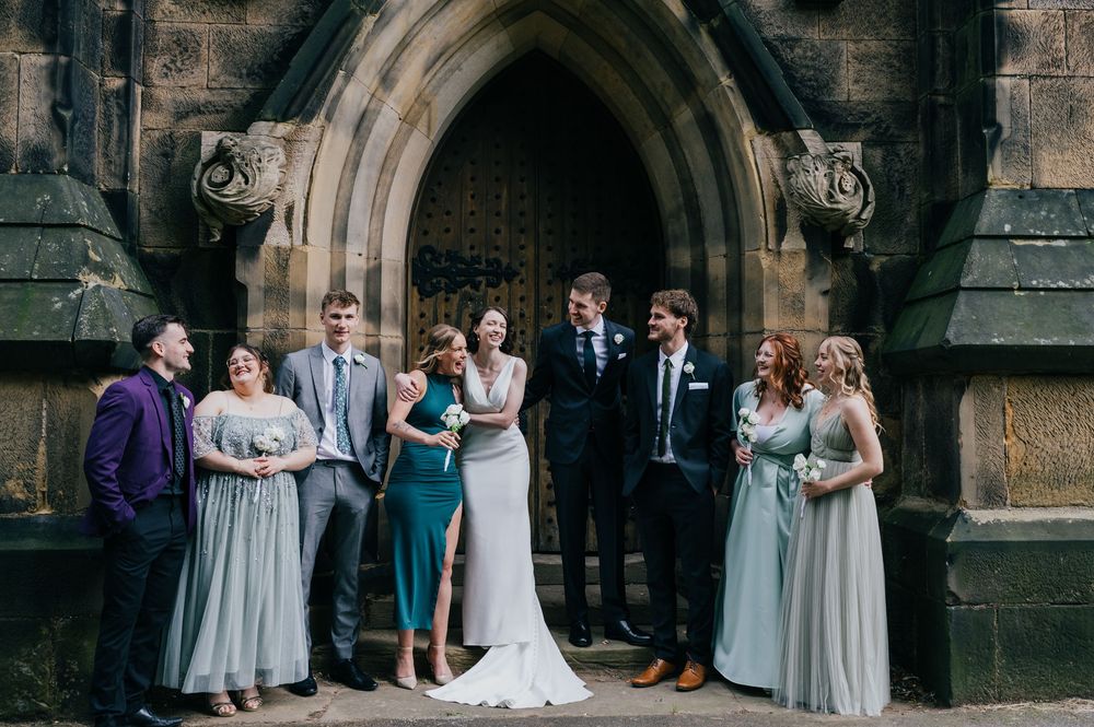 Wedding party members pose together in front of a stone church exterior for group photos.