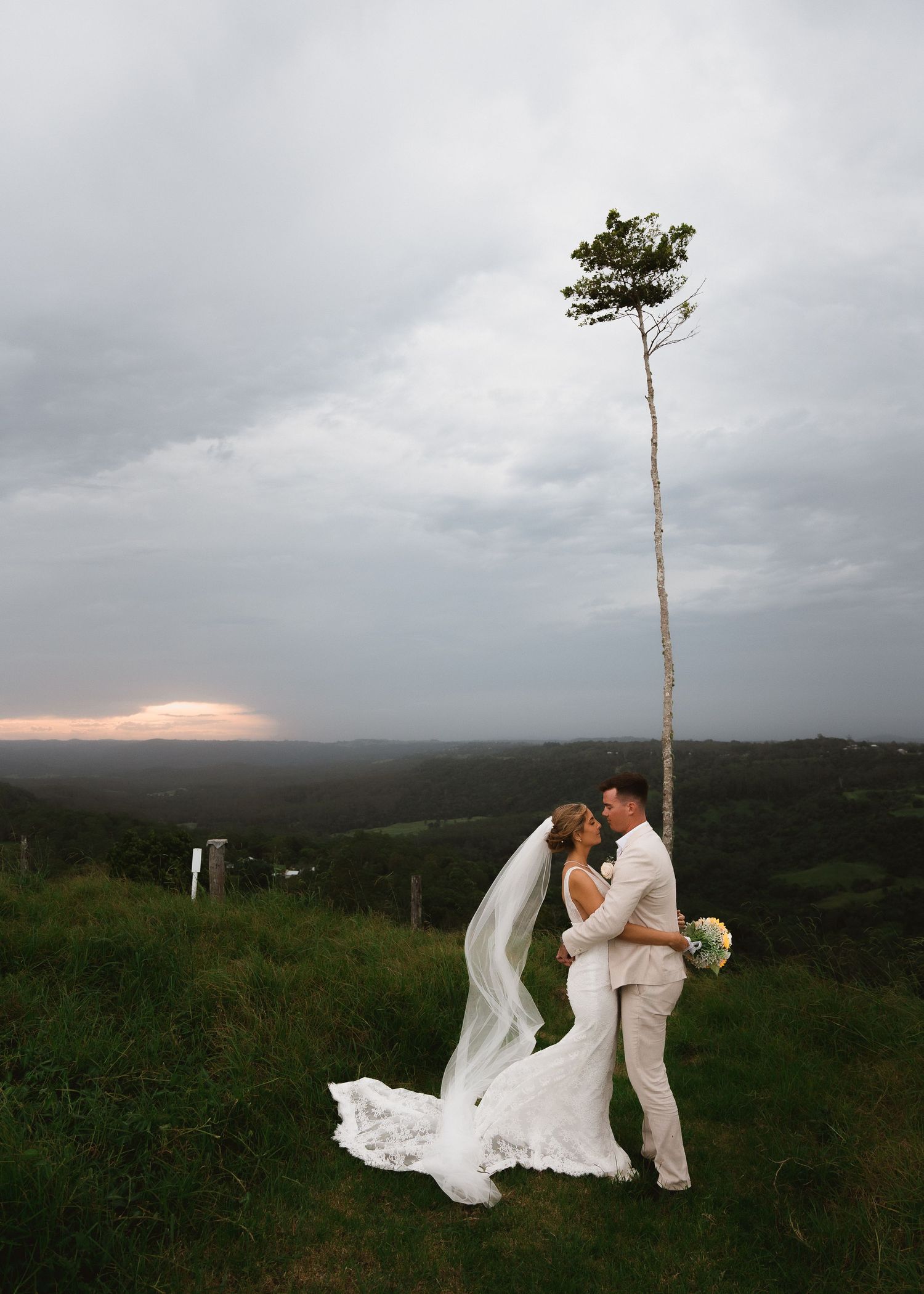 Newlyweds sharing a romantic moment under a lone tall tree against a moody sky at sunset.