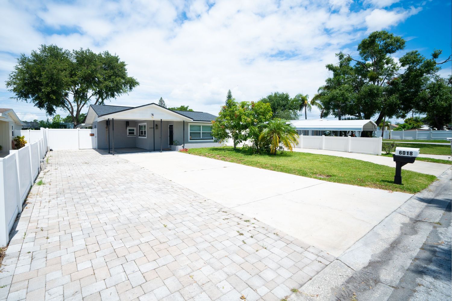 Modern home exterior with paved driveway, white fence, and tropical landscaping under bright blue Florida sky.