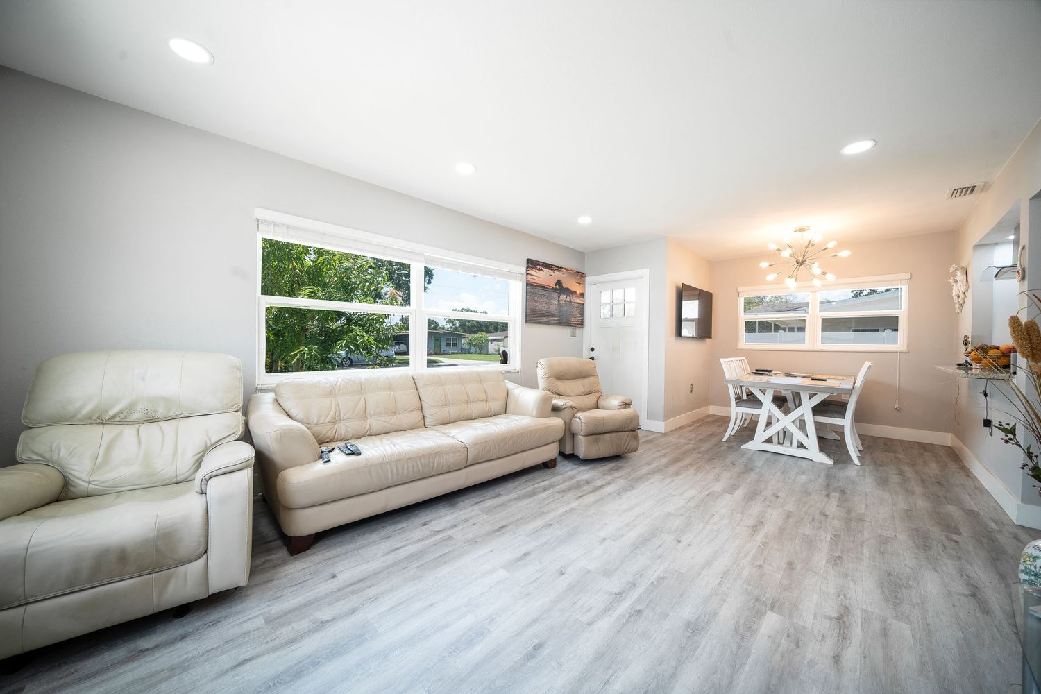 Modern living room with cream leather sofas, light gray wood floors and dining area featuring bright natural lighting.