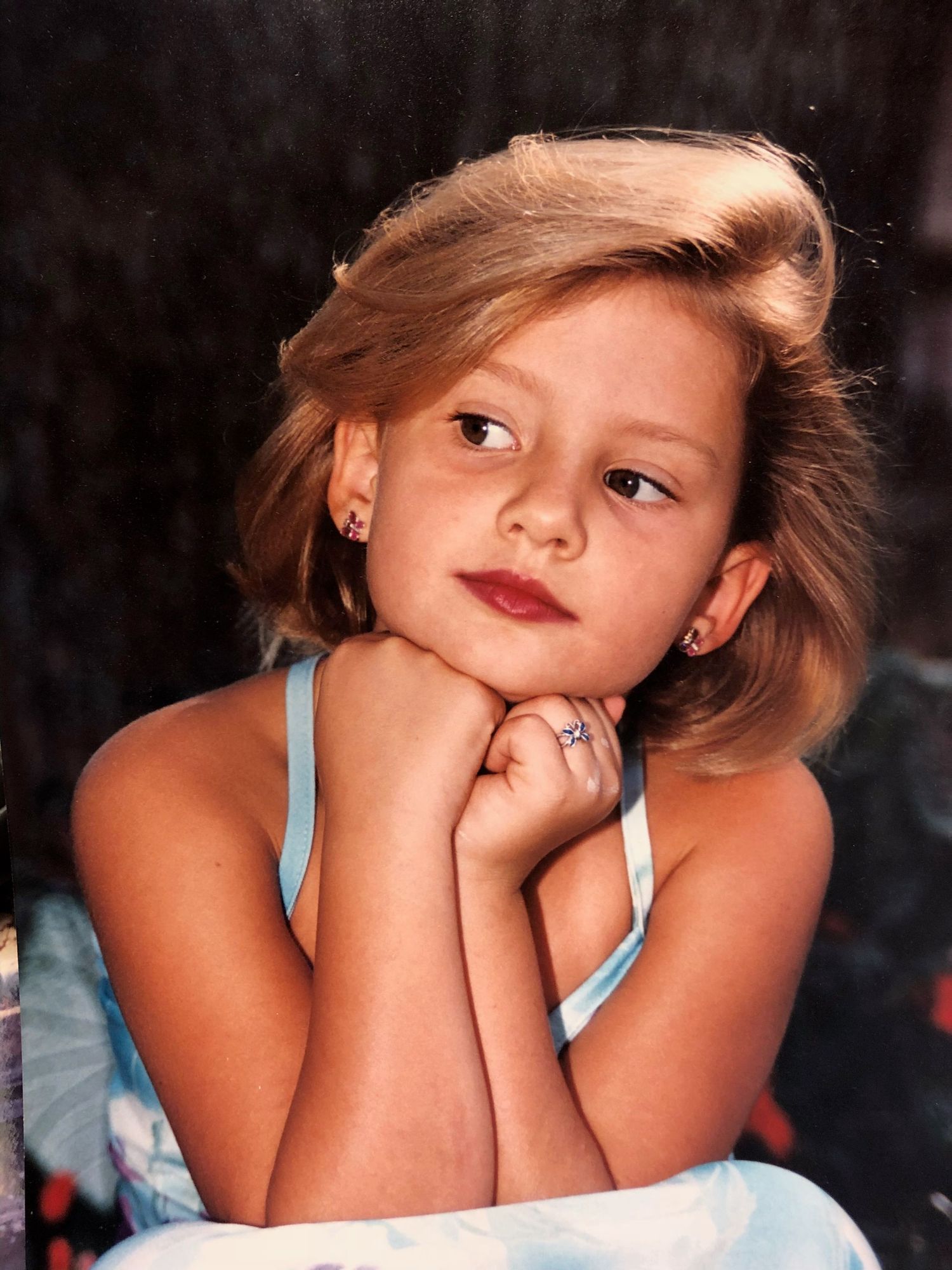 A portrait of a young person in a light blue tank top resting their chin on their hands against a dark background.