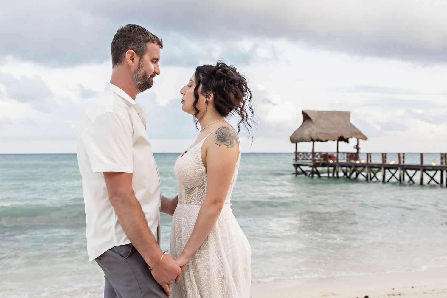 The bride and the groom hold hands near the water, with the wooden pier of The Fives Beach in the background.