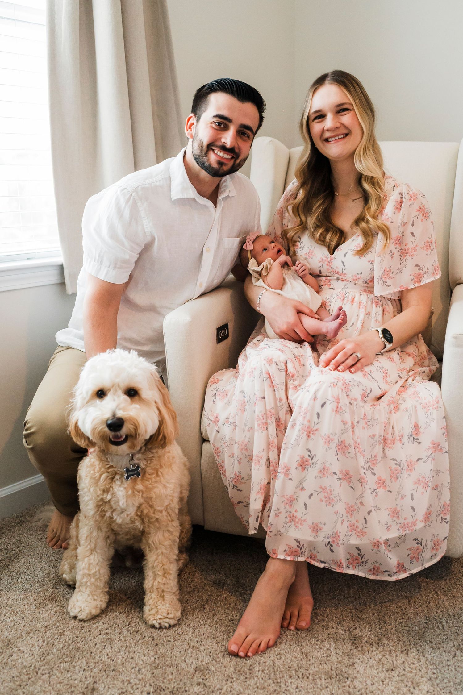 A couple sitting on a bed with their newborn baby and a fluffy goldendoodle dog with pink plush toys.
