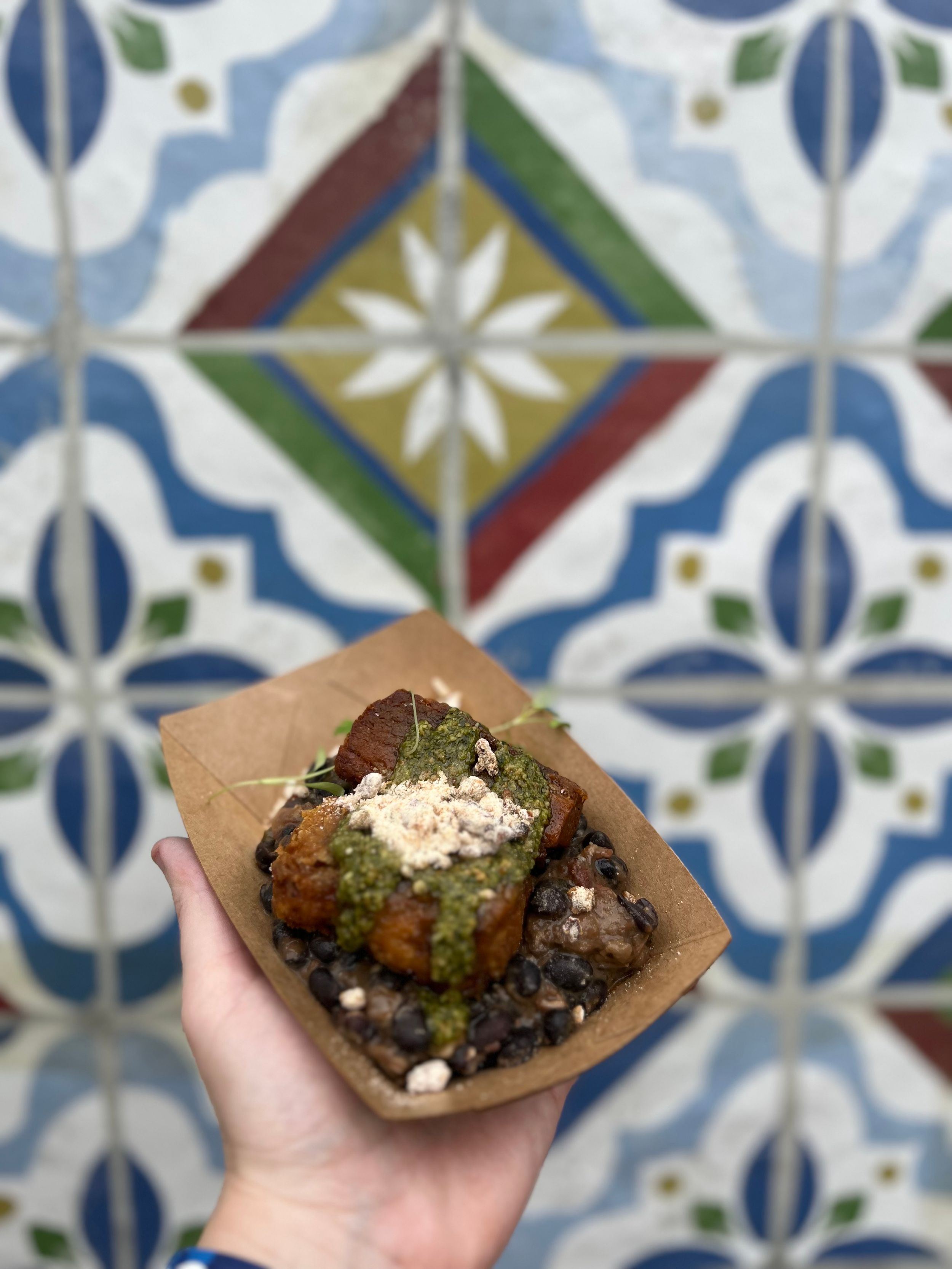 A gourmet street food dish served in brown paper against decorative blue and white tile background.