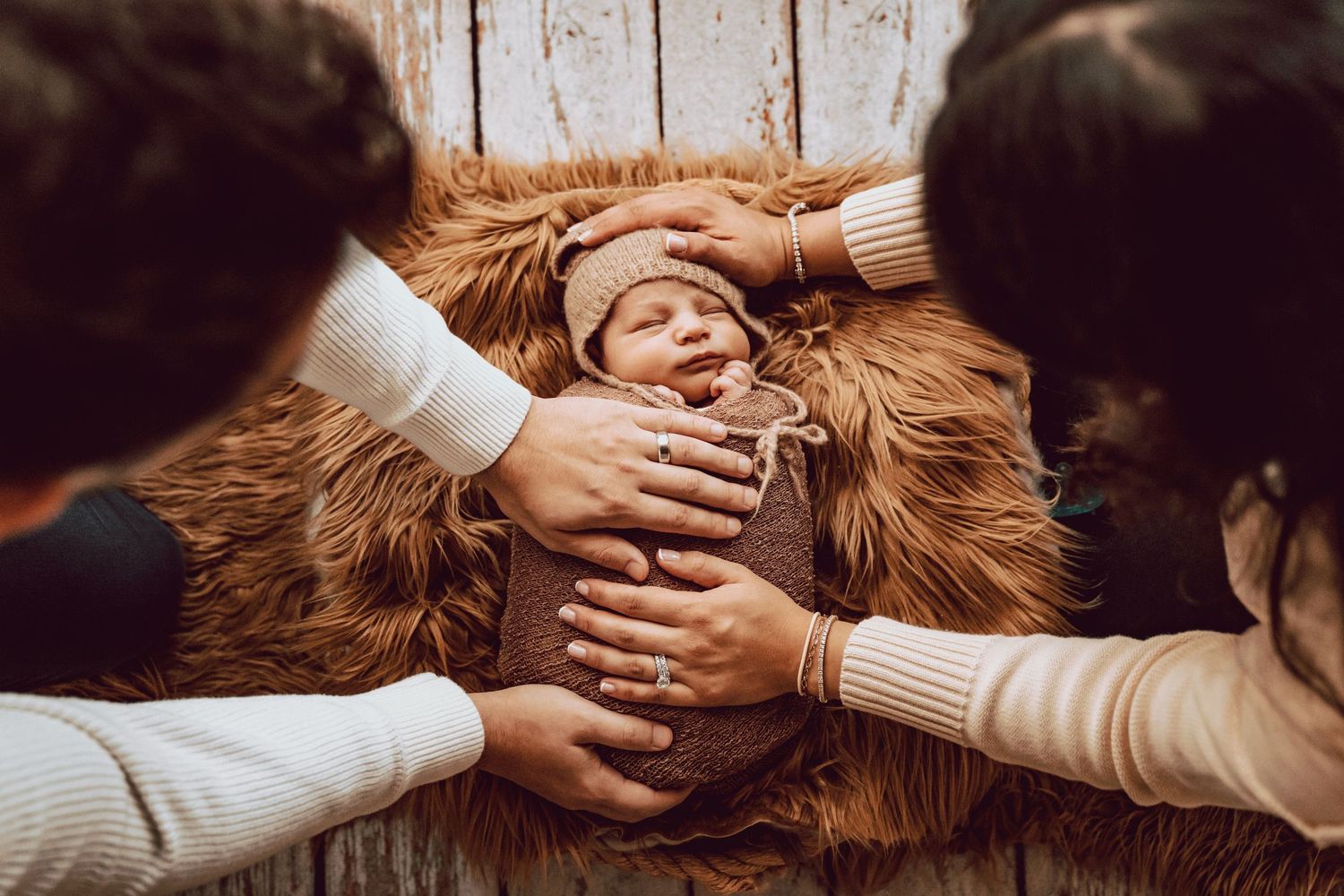 Parents gently cradling their newborn baby wrapped in brown knit on a fluffy fur rug during a newborn photo session.