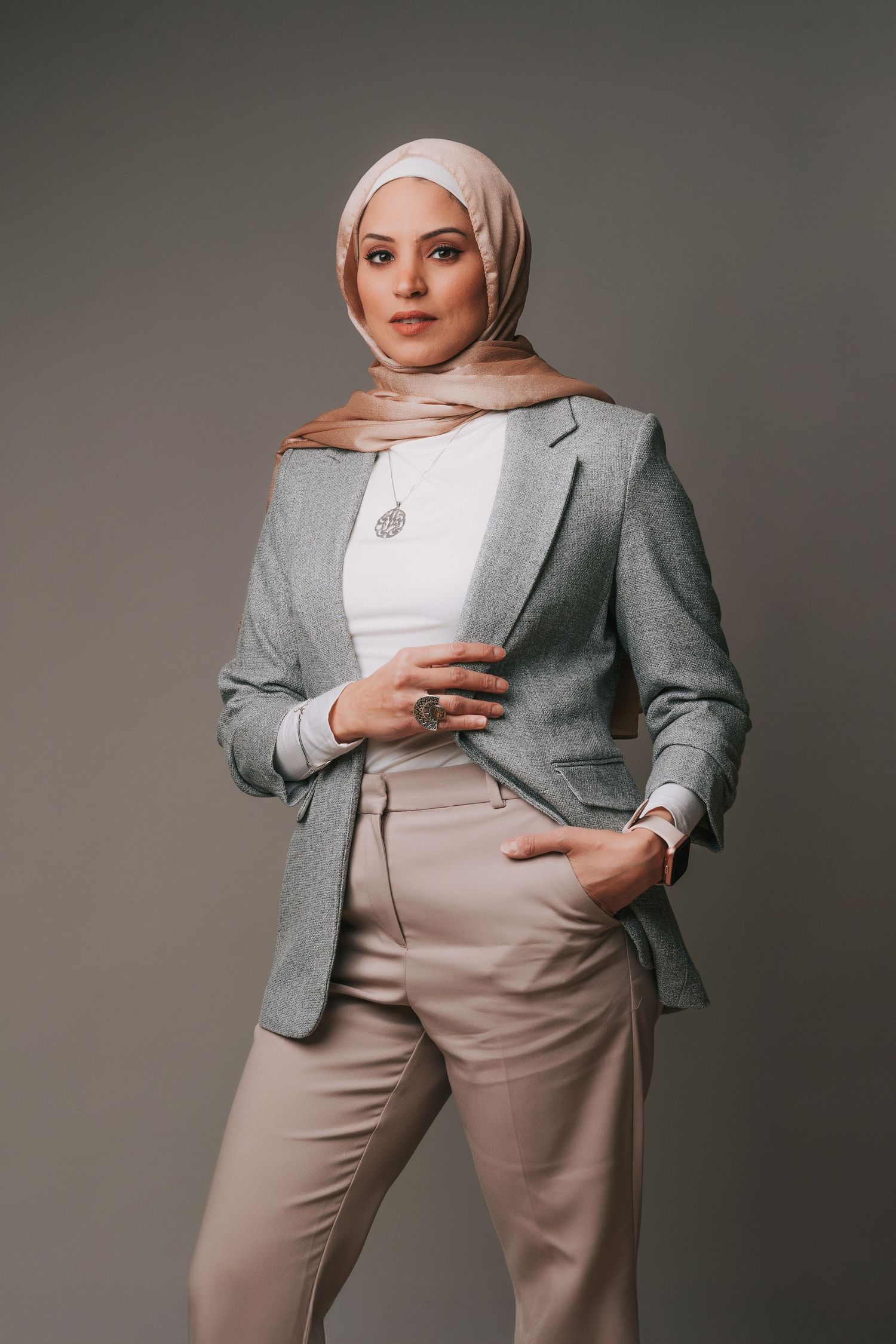 A professional headshot of a woman wearing a light beige hijab, gray blazer, white top, and beige pants standing confidently in studio set