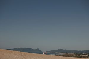 Two people walking along a sandy beach ridge with mountains and blue sky in the distance.