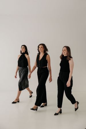 Three women walking in elegant black outfits against a white studio backdrop, showcasing modern minimalist fashion.