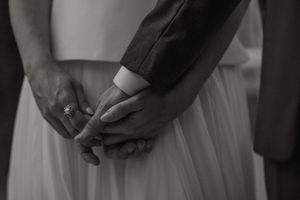 A couple holds hands during their wedding ceremony with white dresses and decor visible in the background.