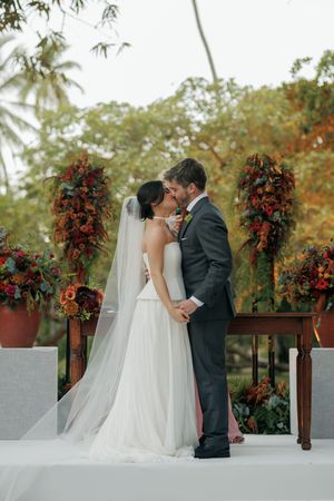 A romantic wedding scene with red floral arrangements and palm trees in the background on an outdoor terrace.