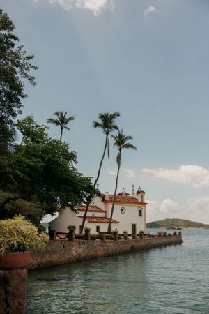 A small white church with Spanish-style architecture sits on a rocky peninsula surrounded by palm trees and ocean waters.