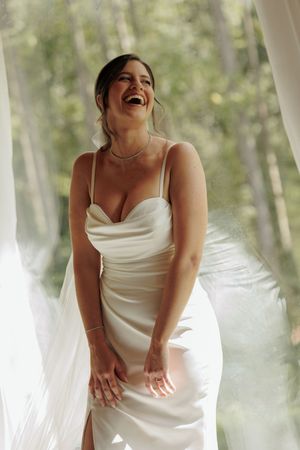 A bride in a white satin dress laughs joyfully during a photo session in a dreamy forest setting.