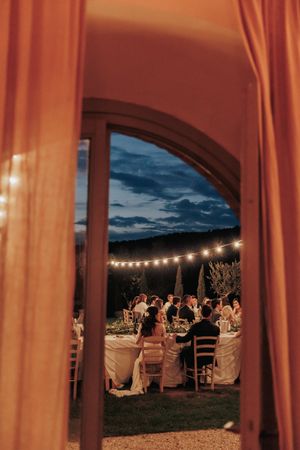 View through curtains of an outdoor evening dinner party with string lights and guests seated at long tables under dark clouds.
