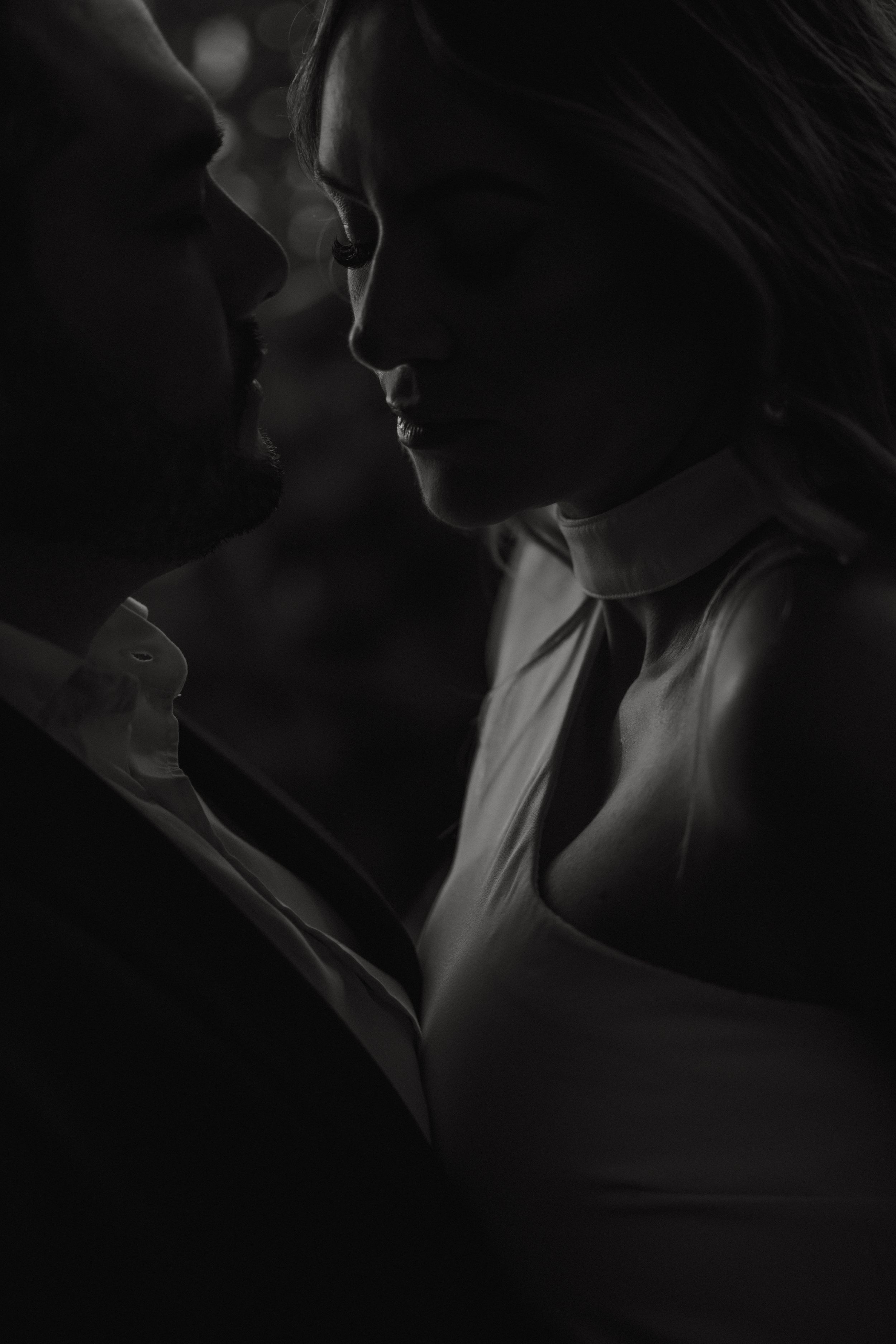 A romantic black and white portrait of a couple in intimate proximity, captured in dramatic shadows and low light.
