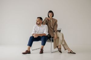 Two people in modern minimalist attire pose together against a white backdrop in a studio setting.
