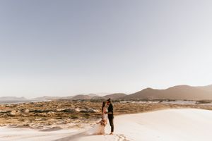 A couple poses in a desert setting during a golden hour photoshoot as the sun casts warm light across the sandy landscape.