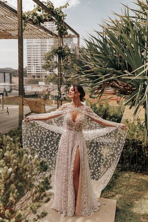 A sheer lace wedding dress with flowing sleeves is displayed in a tropical garden setting with palm trees and city views.
