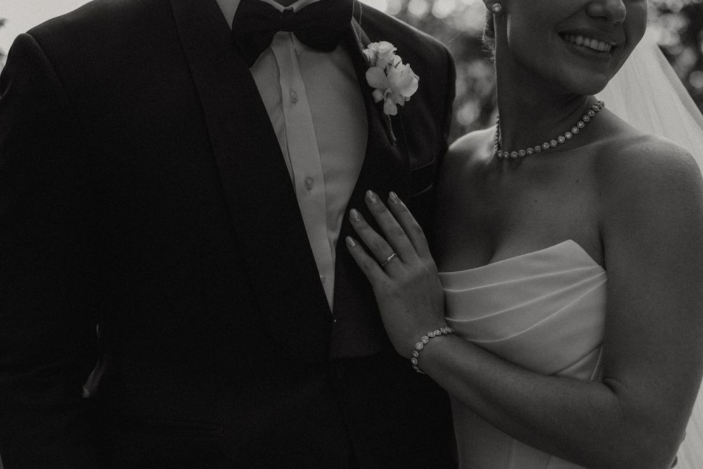 Black and white close-up wedding photo showing couple's formal attire with tuxedo, pearl necklace and strapless dress.