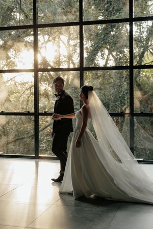 A silhouetted bride and groom stand before floor-to-ceiling windows with sunlight streaming through trees outside.