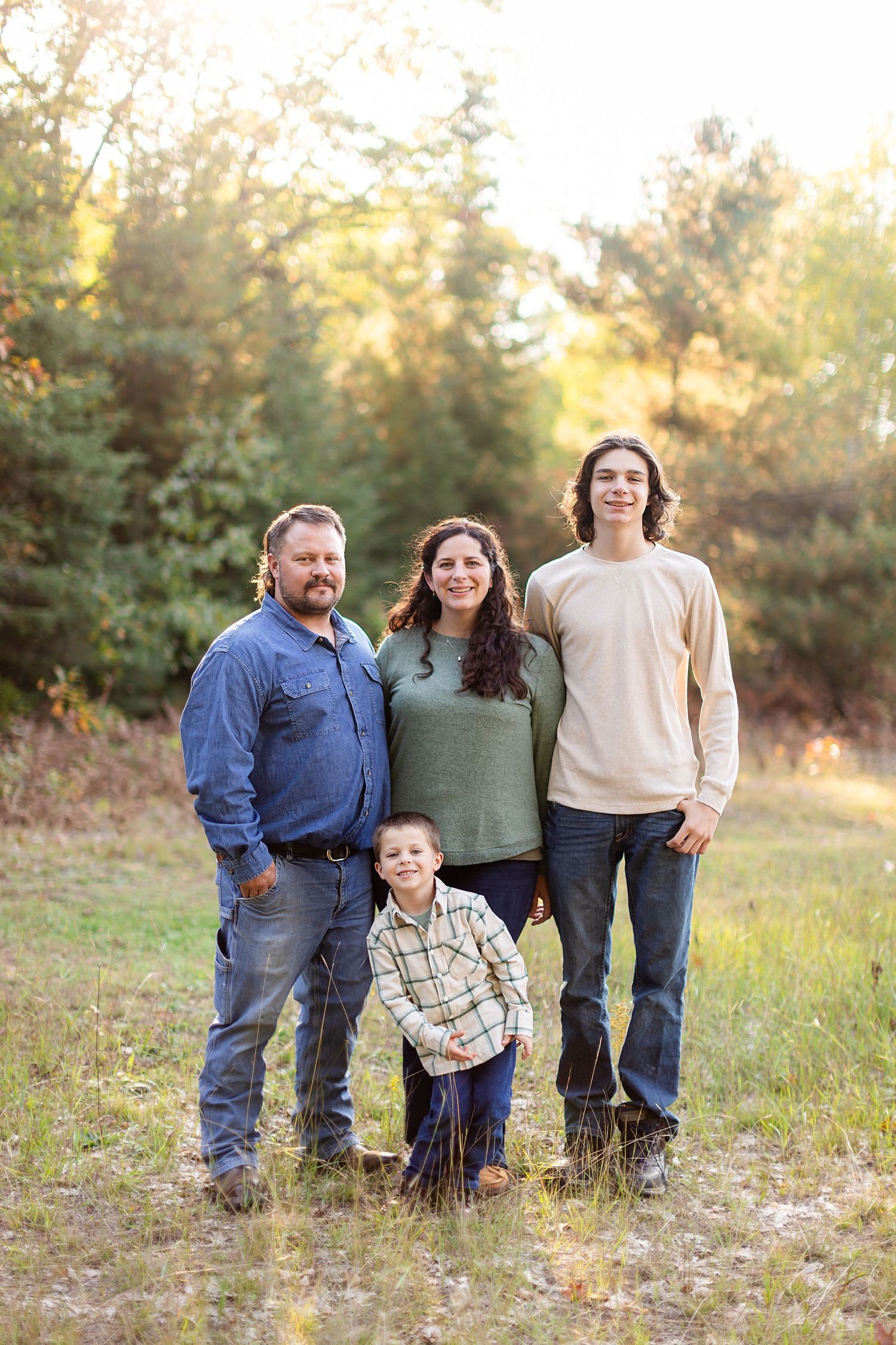 A family of four poses together outdoors in a field with autumn trees and golden light in the background.