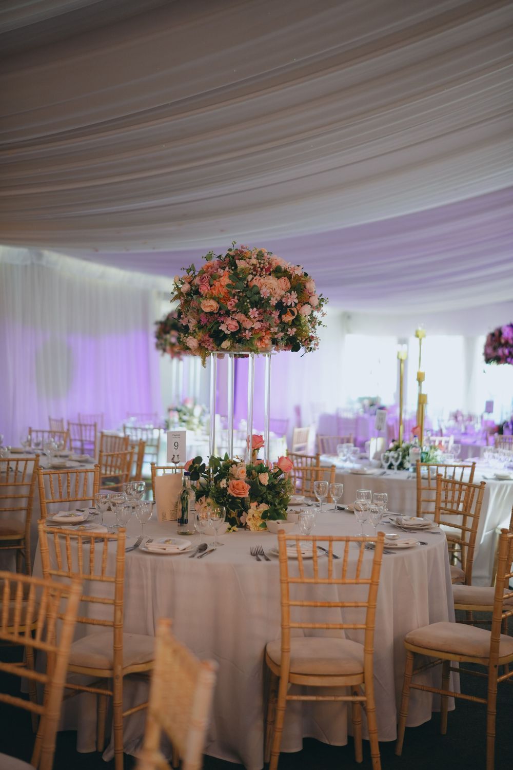 Elegant wedding reception setup with tall coral floral centerpieces and chiavari chairs in a white draped tent venue.