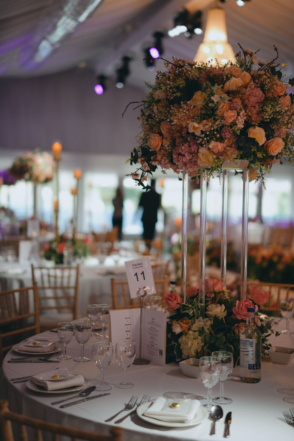 Elegant wedding reception table setting with tall floral centerpiece featuring peach and coral blooms in a glass vase.
