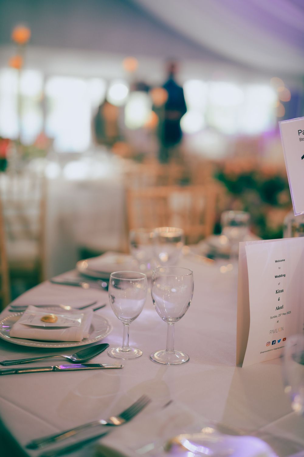 Elegant wedding reception table setting with white linens, wine glasses and menu cards in soft lighting.