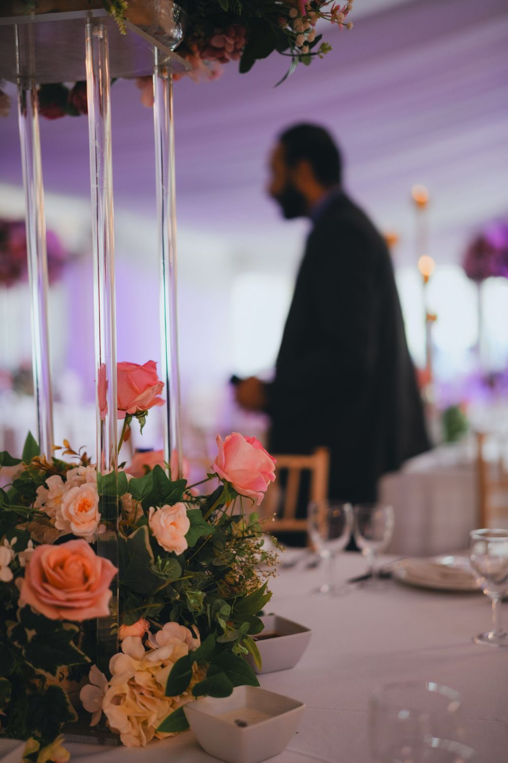 Silhouetted figure walks past romantic table decorations with coral roses and tall glass vase centerpieces.