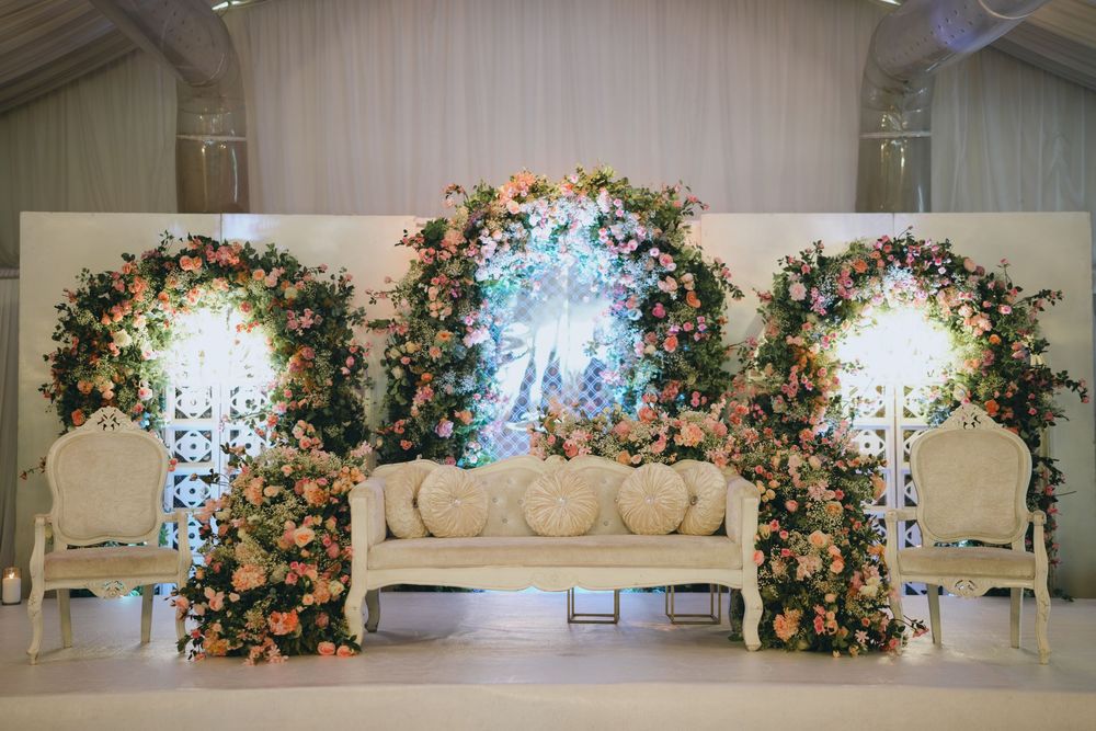 Elegant wedding stage setup with floral arches, crystal chandeliers and cream-colored seating against white backdrop.