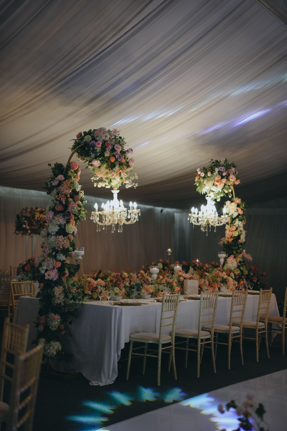 Elegant wedding reception venue with crystal chandeliers and floral arches adorning a long white dining table.