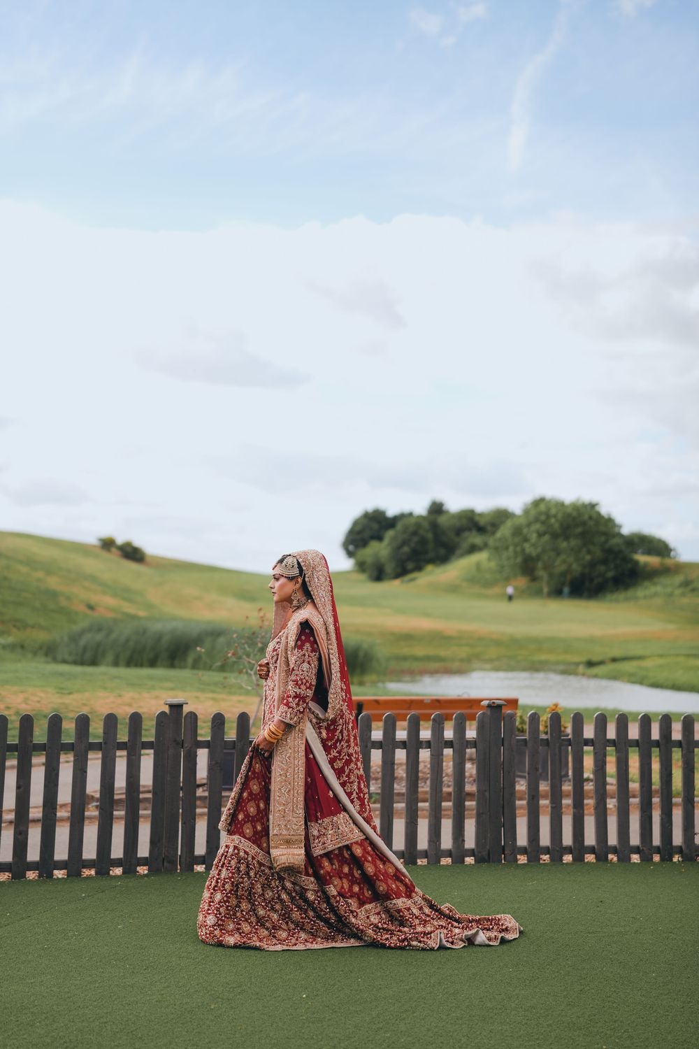 Traditional Indian bride in red lehenga standing on a golf course with scenic rolling hills.