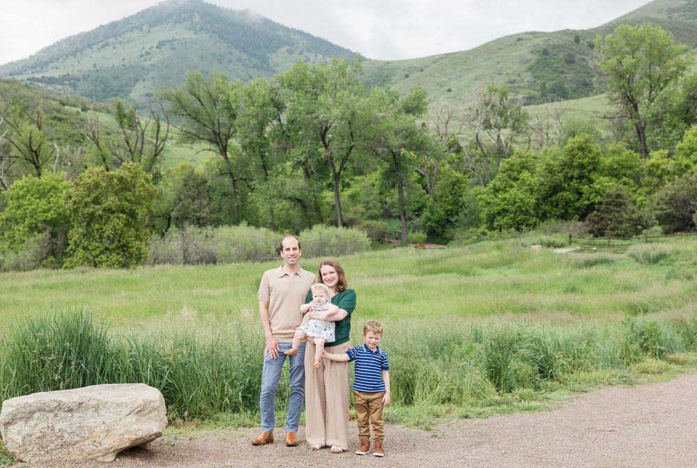 Small family enjoys nature together while standing on dirt path near meadow with green mountains in background.