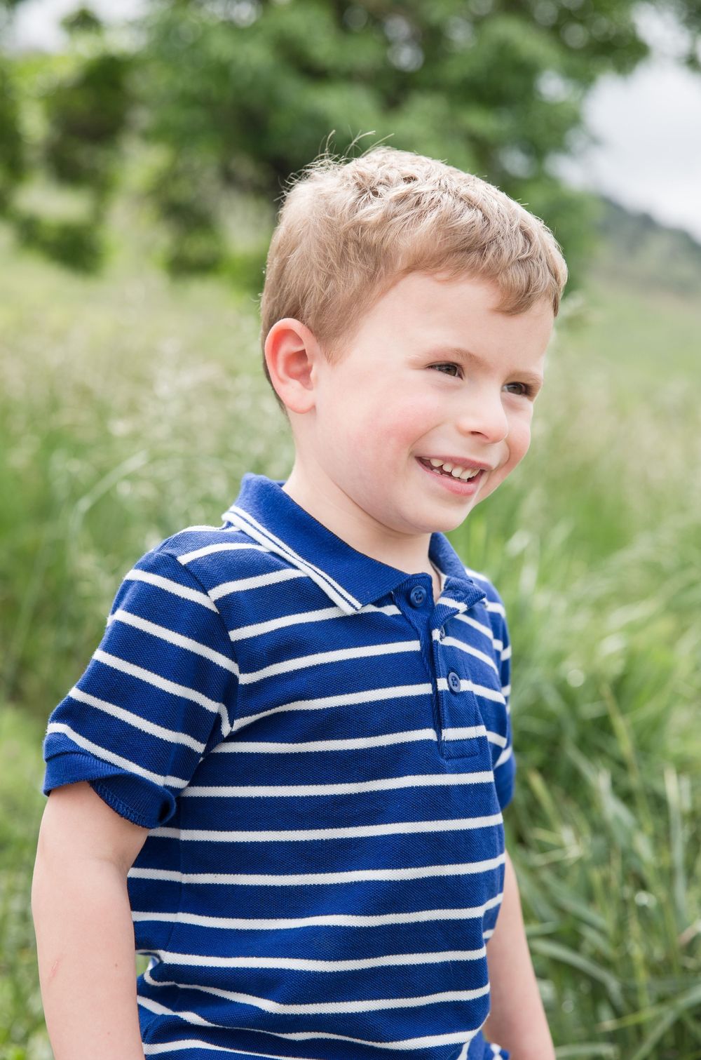 A smiling child wearing a blue and white striped polo shirt stands in a grassy meadow.