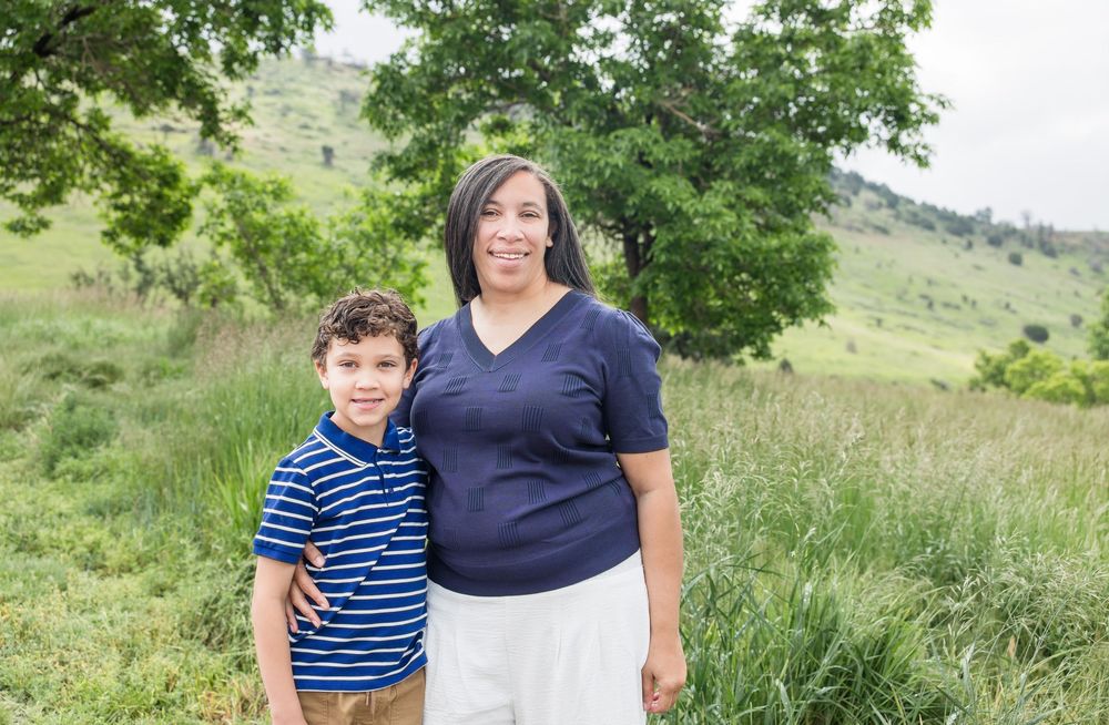 Two people stand together in a grassy field on a summer day with green hills and trees in the background.