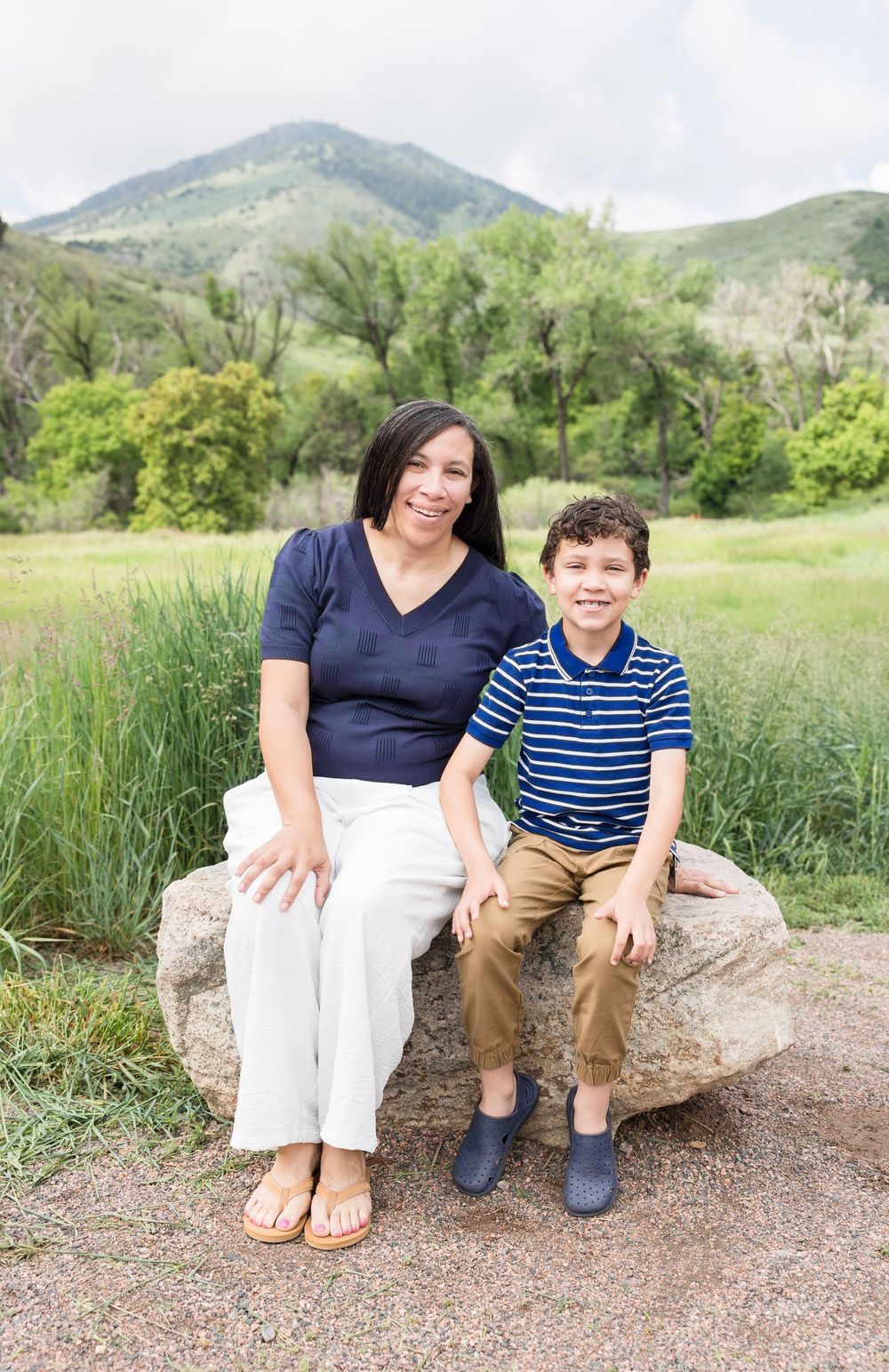 Two people sit together on a large rock in a scenic mountain landscape with green hills and vegetation.