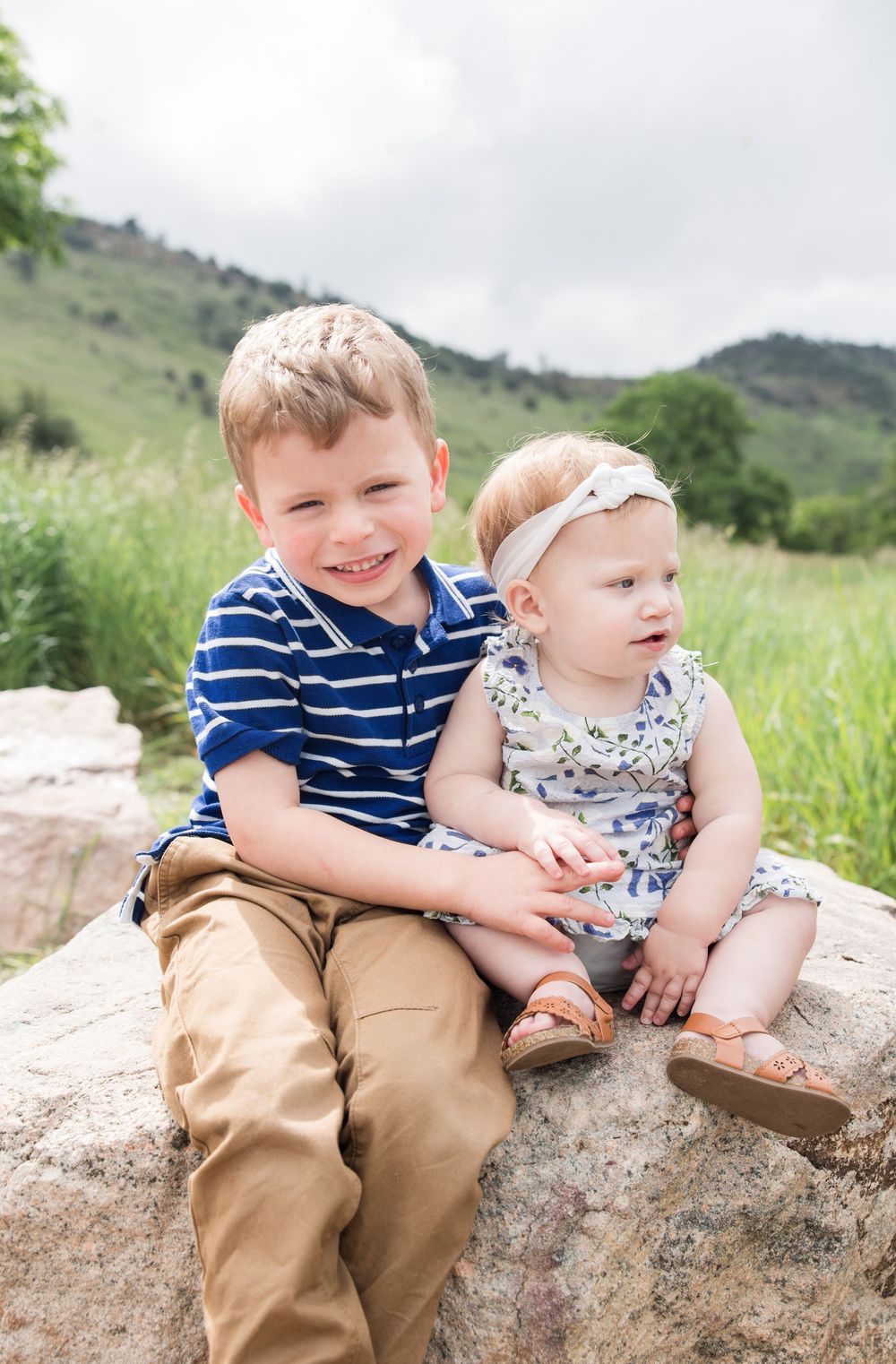 Two children sit together on a large rock in a grassy mountain landscape on a cloudy day.