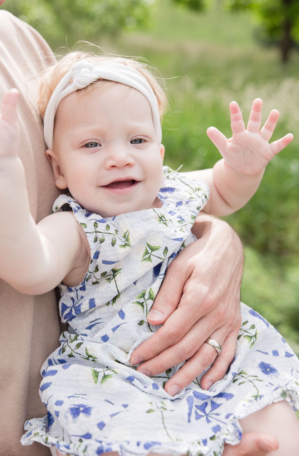 Baby in a floral dress waves while wearing a white headband in an outdoor garden setting.