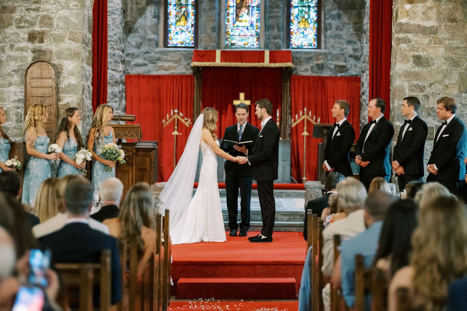 A wedding ceremony takes place inside a historic stone church with stained glass windows and red carpeted altar area.