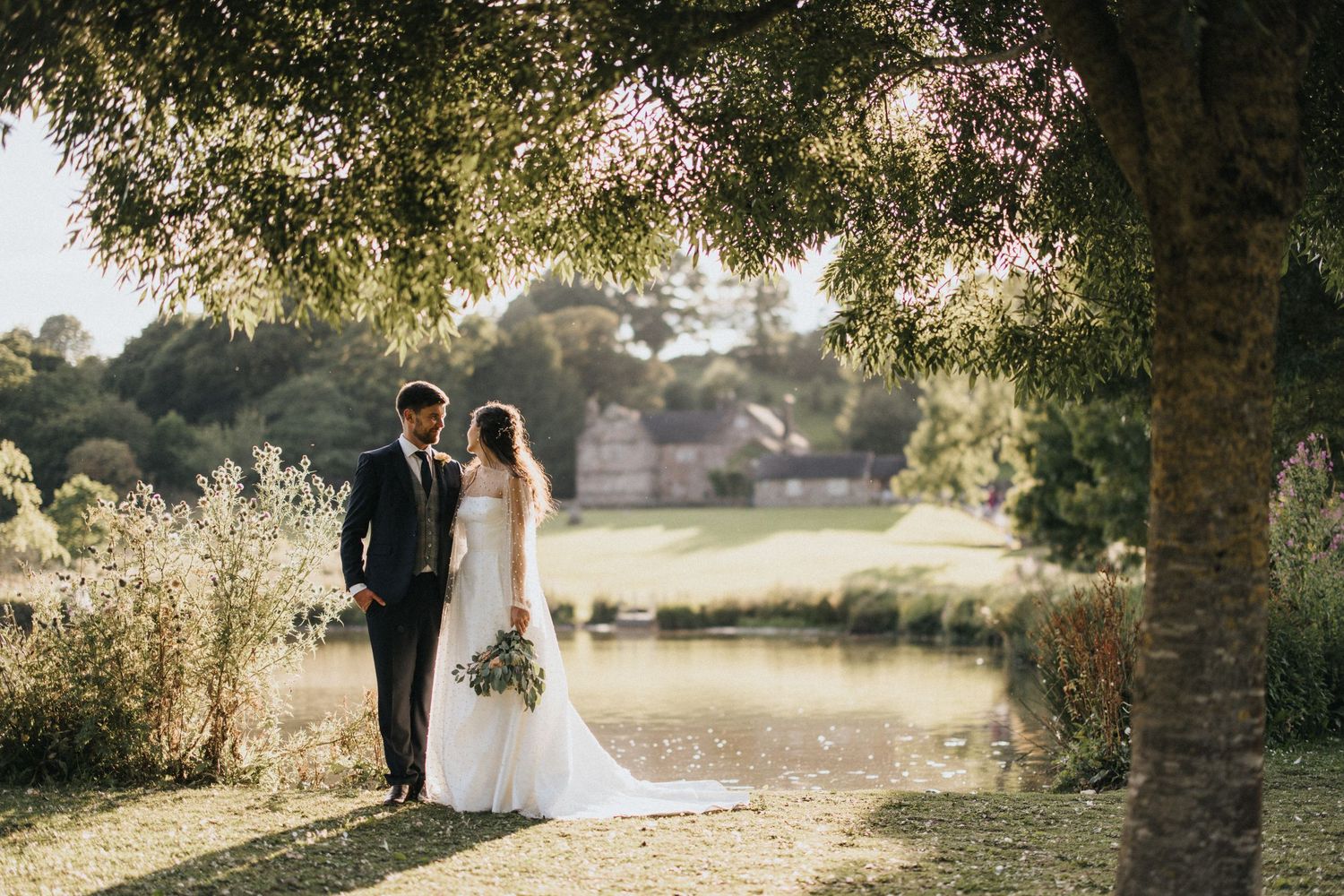 Wedding couple walks hand in hand through sun-dappled trees beside a tranquil lake at golden hour.