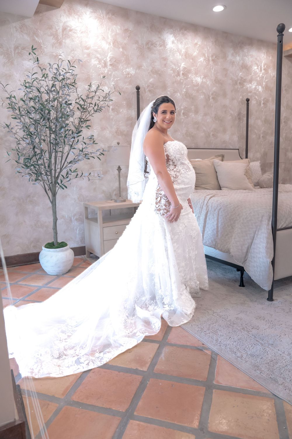 A bride in a stunning lace wedding gown with long train poses in a bedroom with terra cotta floors and potted greenery.
