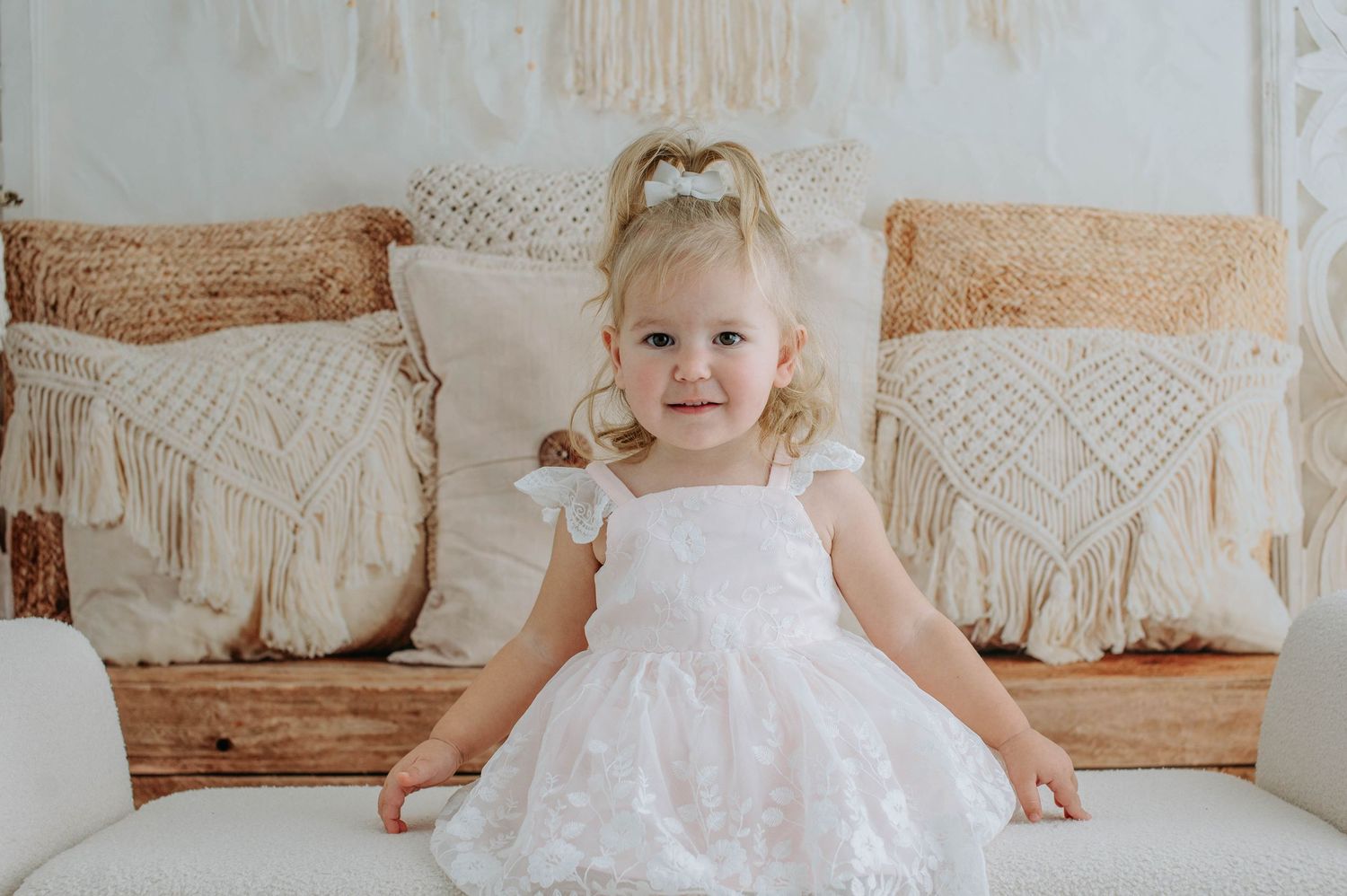 A white lace dress adorns a small figure seated on a couch with boho macrame pillows against a light wall.