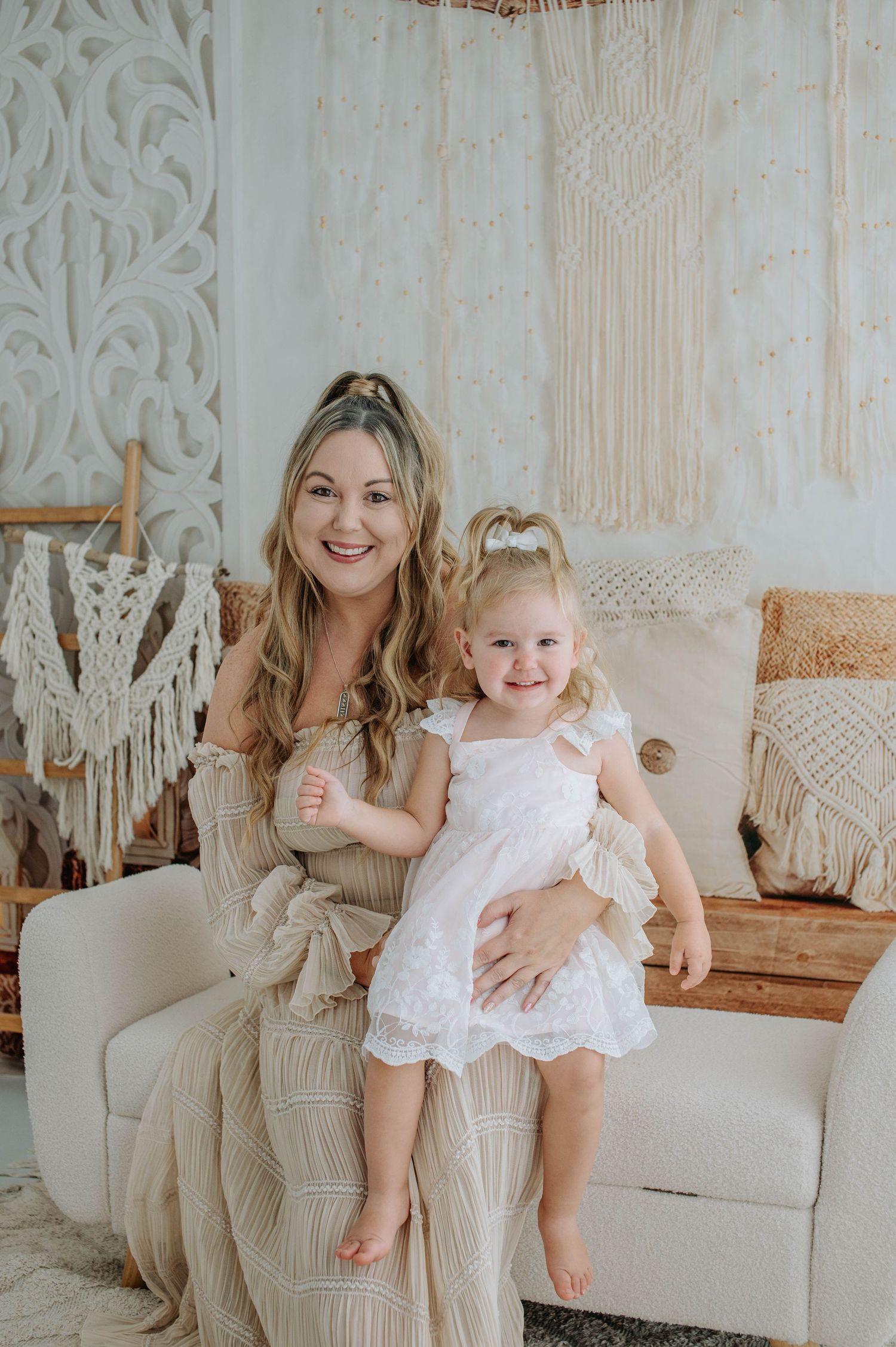 Mother and baby daughter in white dresses sitting together on beige couch with macrame wall decor background.