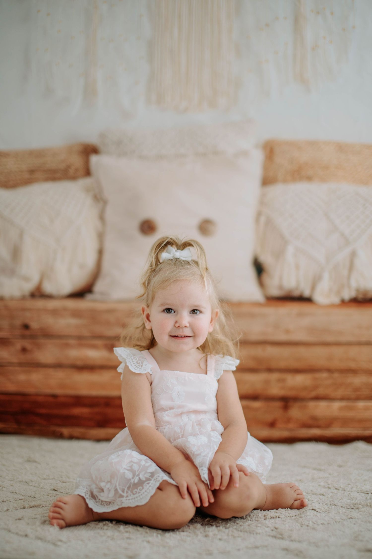 A toddler wearing a white lace dress sits on a carpet against rustic wooden bench with decorative pillows.
