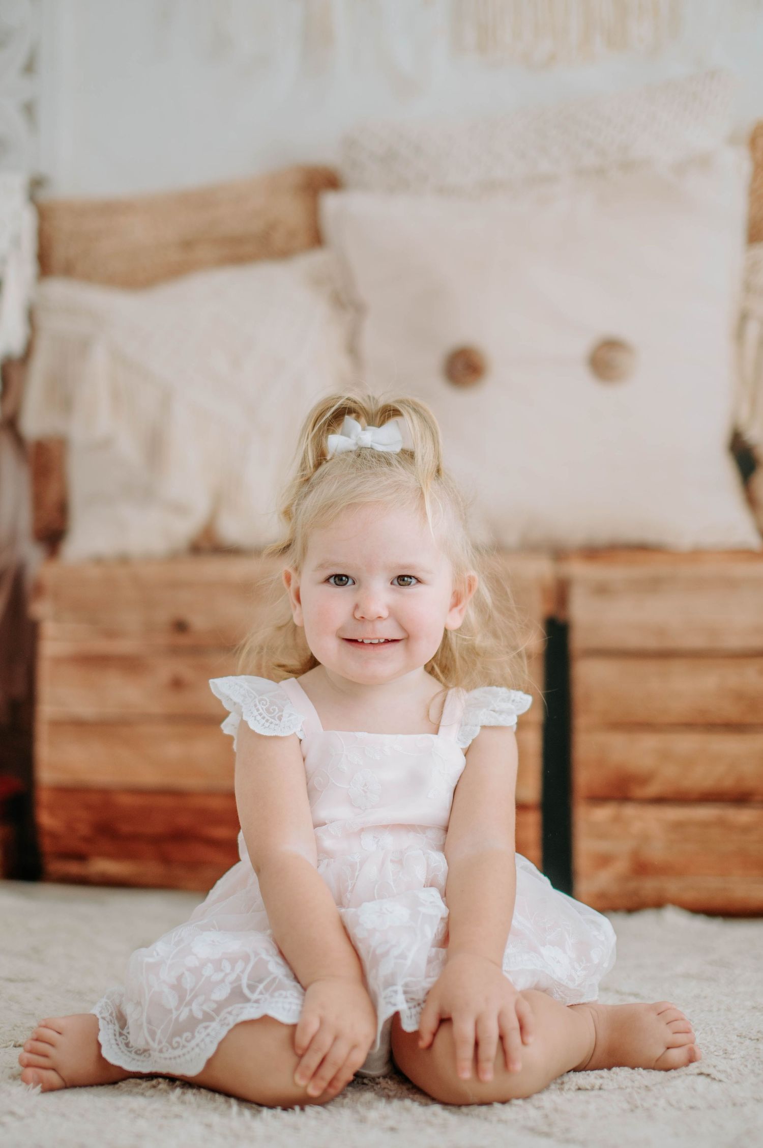 A toddler in a white lace dress sits cross-legged on a white carpet with rustic wood furniture behind.