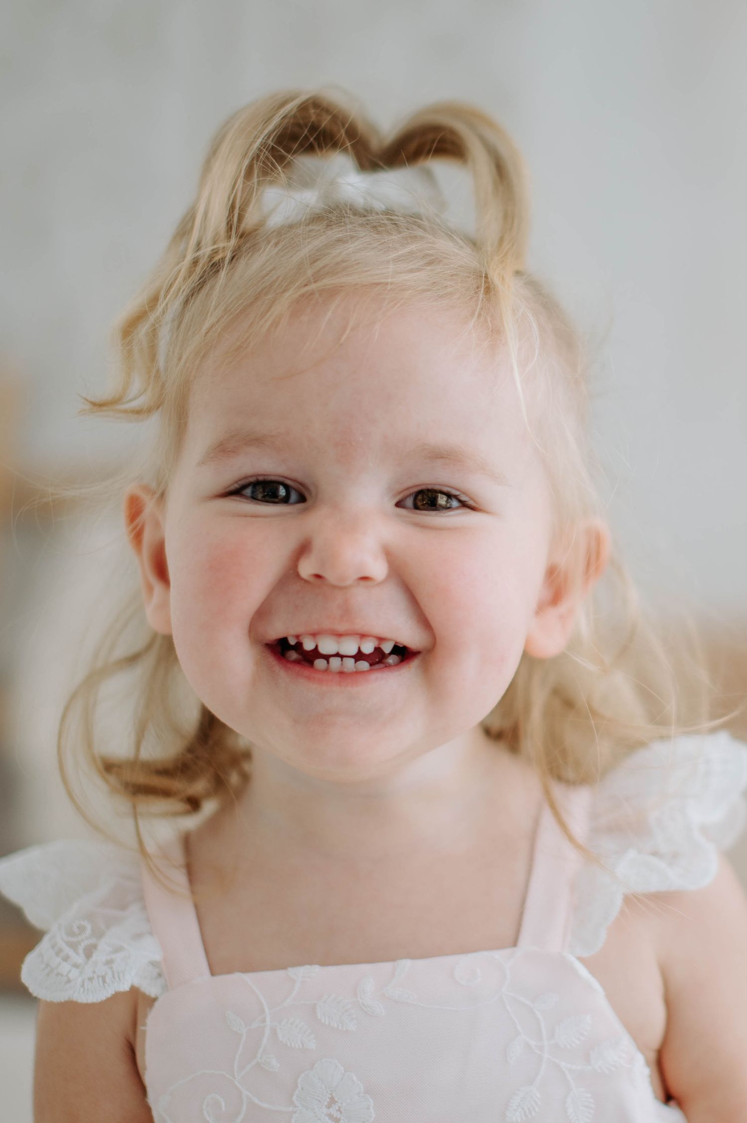 A joyful toddler in a white lace dress smiles brightly against a light background.