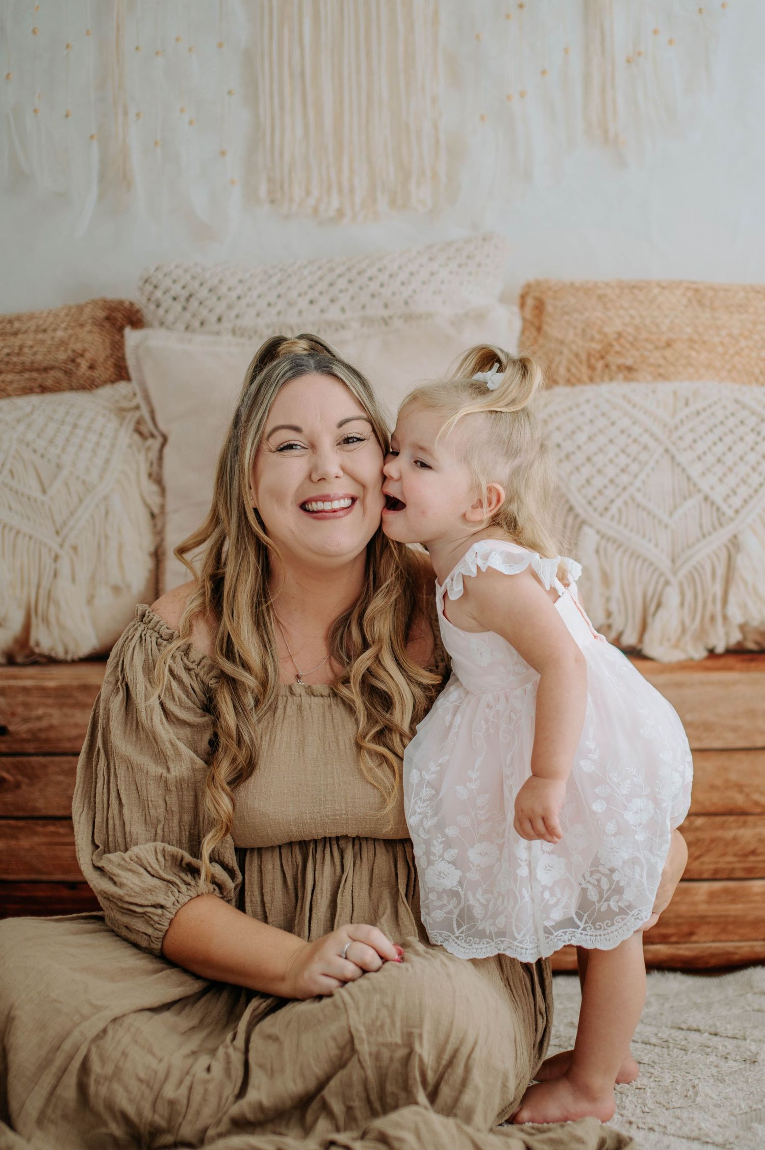 A tender moment captured between a woman in a beige dress and a child in white lace dress sharing smiles and snuggles.
