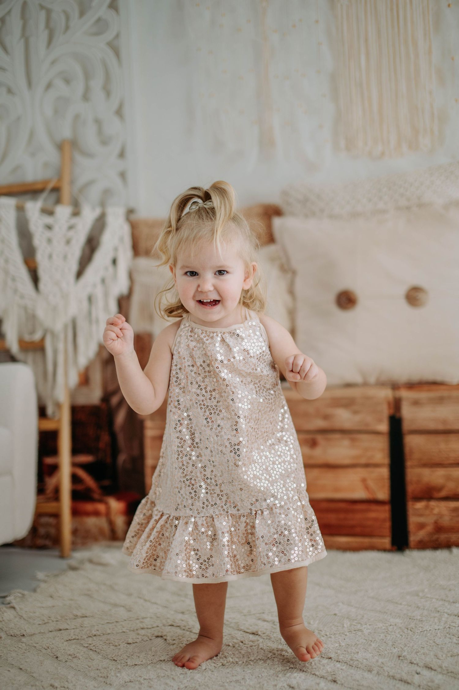 A toddler wearing a shimmering champagne dress stands barefoot on a carpet with decorative backdrop.